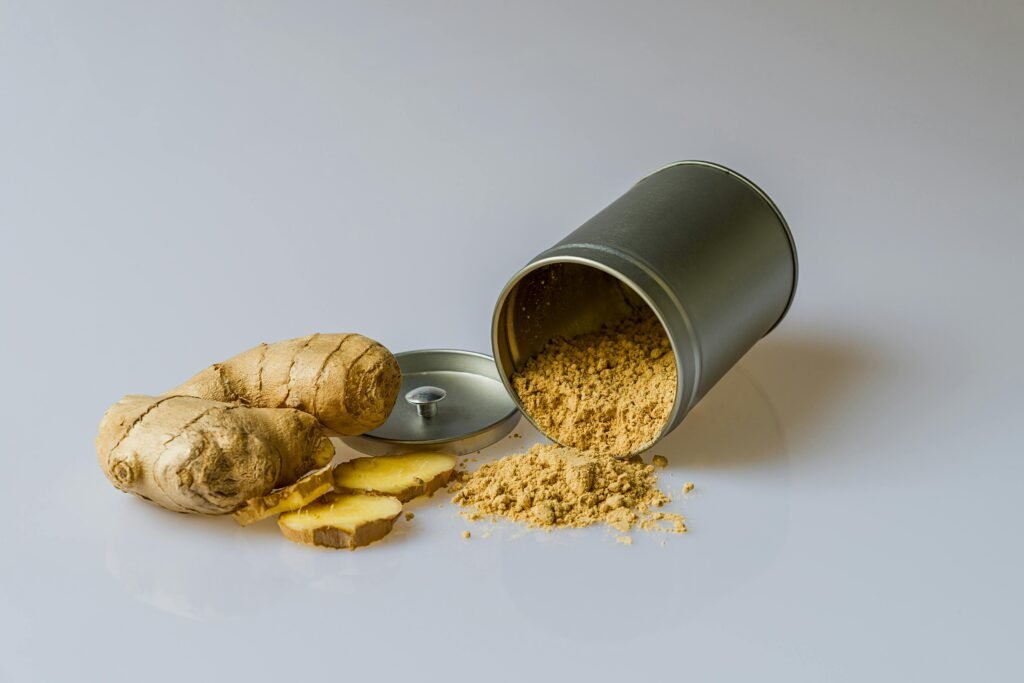A fresh ginger root with several slices cut, placed next to an open metal container spilling ginger powder onto a clean white surface. The image highlights ginger health benefits by showcasing both raw and powdered forms of the spice.