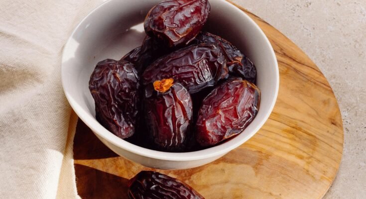 A white ceramic bowl filled with fresh, dark brown dates sits on a round wooden board, with one date resting outside the bowl. The natural lighting highlights the wrinkled texture of the dates. A beige cloth is draped in the background. The image visually represents the benefits of dates, showcasing their rich, nutritious appearance.