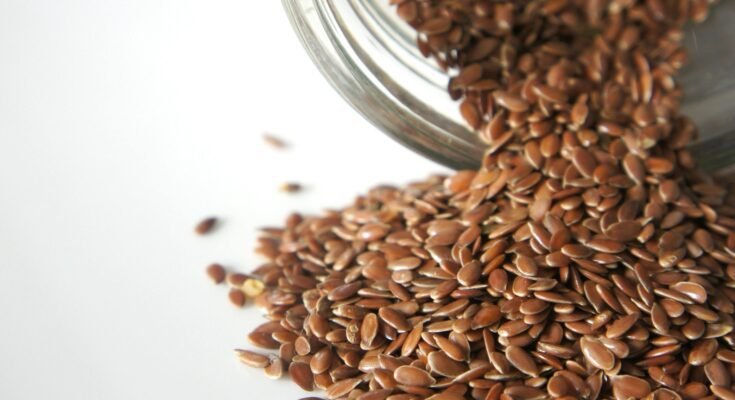 A glass jar tipped over, spilling brown flaxseeds onto a white surface. Flaxseeds are known for their rich nutritional profile, including high fiber, omega-3 fatty acids, and antioxidants. The benefits of flaxseeds include improved digestion, heart health support, and potential weight management.